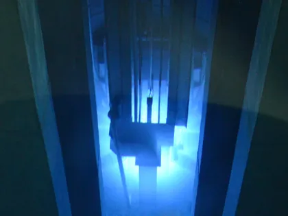 Looking down a narrow shaft into a reactor pool, the water around the submerged assembly lit blue by Cherenkov radiation.